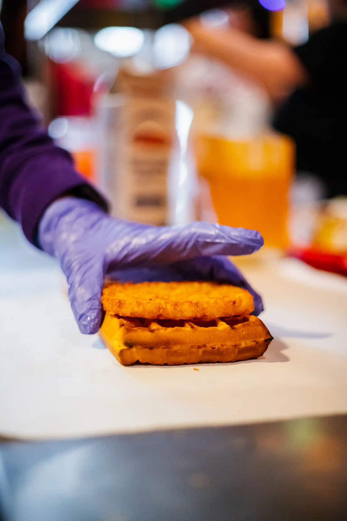 Hand wearing a purple glove holding a waffle and a hashbrown on a counter with a blurred background.