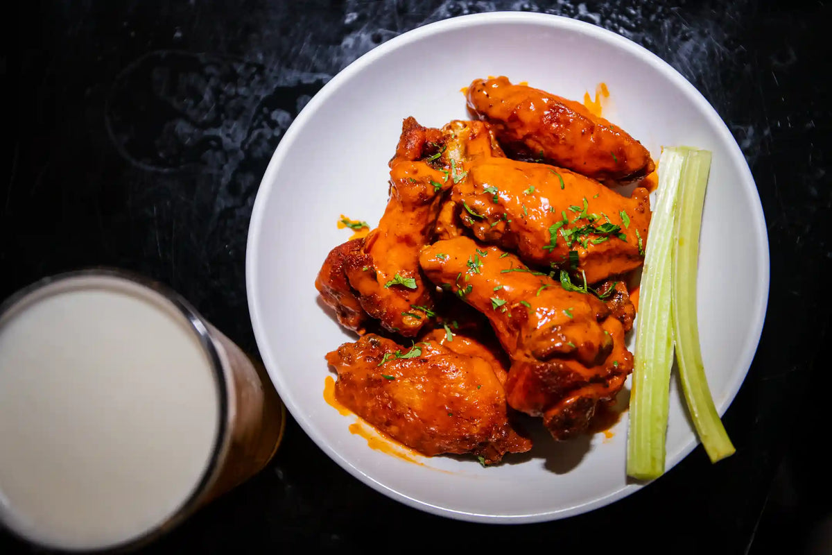 Baked chicken wings on a white plate with celery sticks and a glass of beer on a dark surface