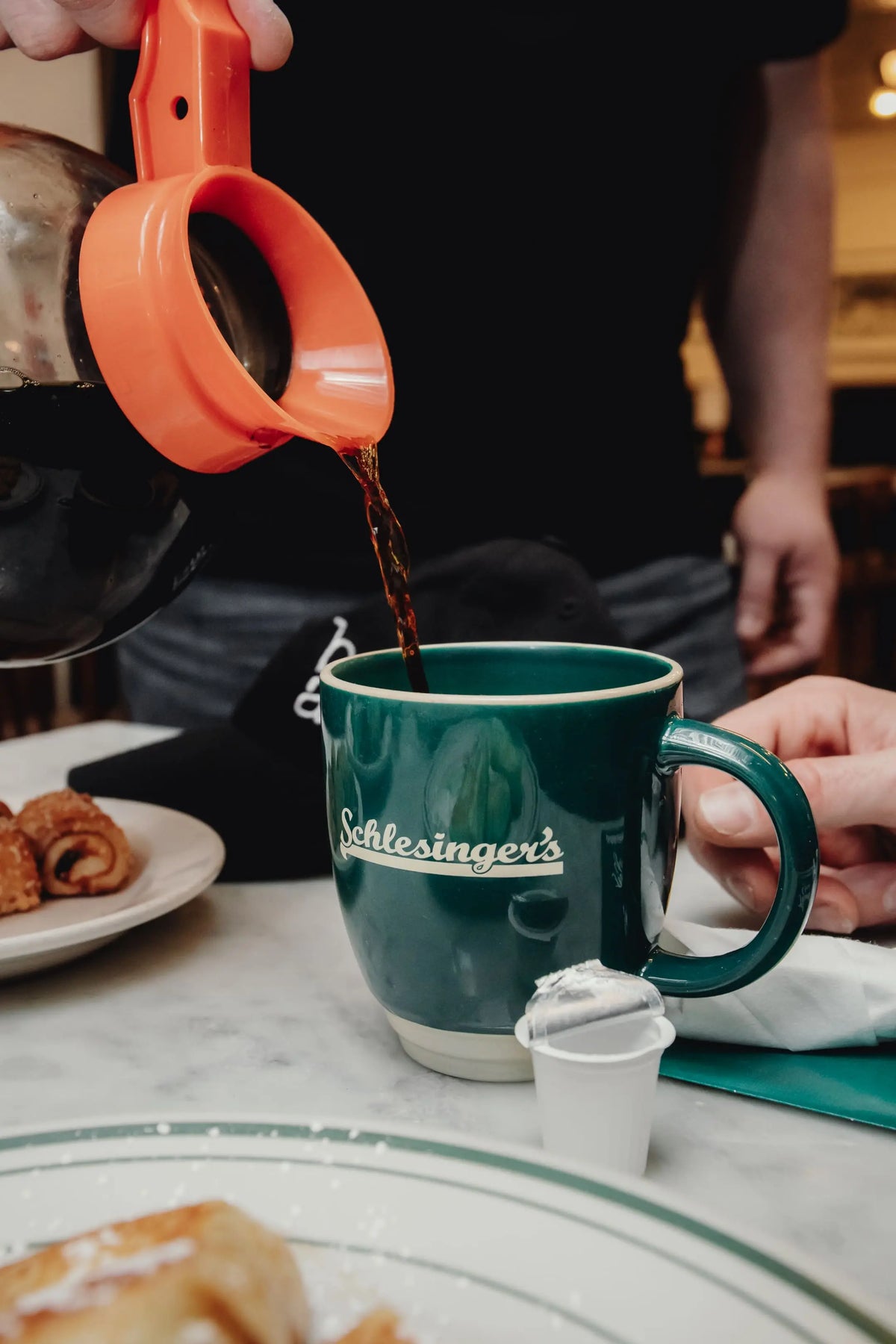 Person pouring coffee from a coffee maker into a green mug with 'Schlesinger's' branding, on a table with pastries.