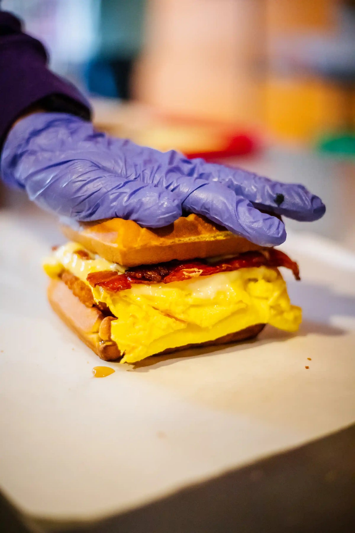 Hand wearing a purple glove holding a breakfast waffle sandwich with yellow cheese on a white surface.