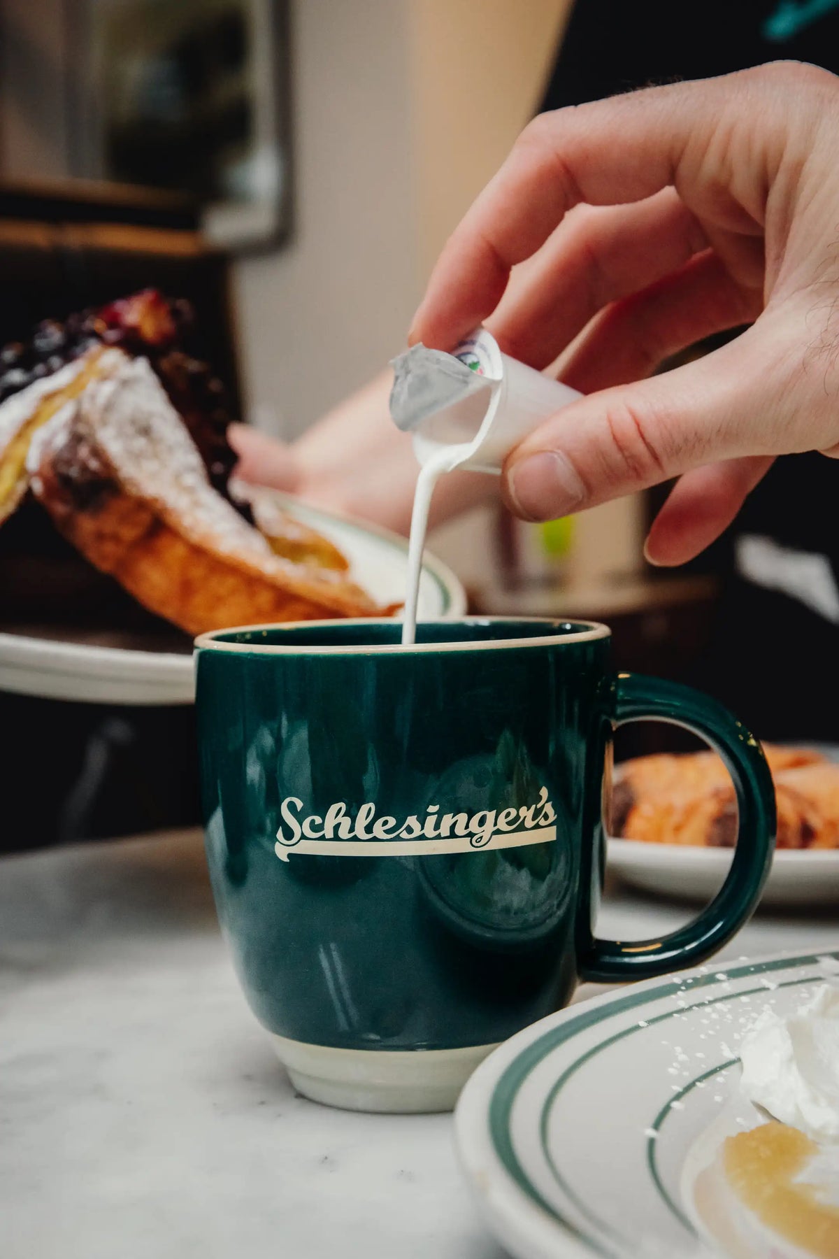 Person pouring creamer into a Schlesinger's mug with pastries in the background