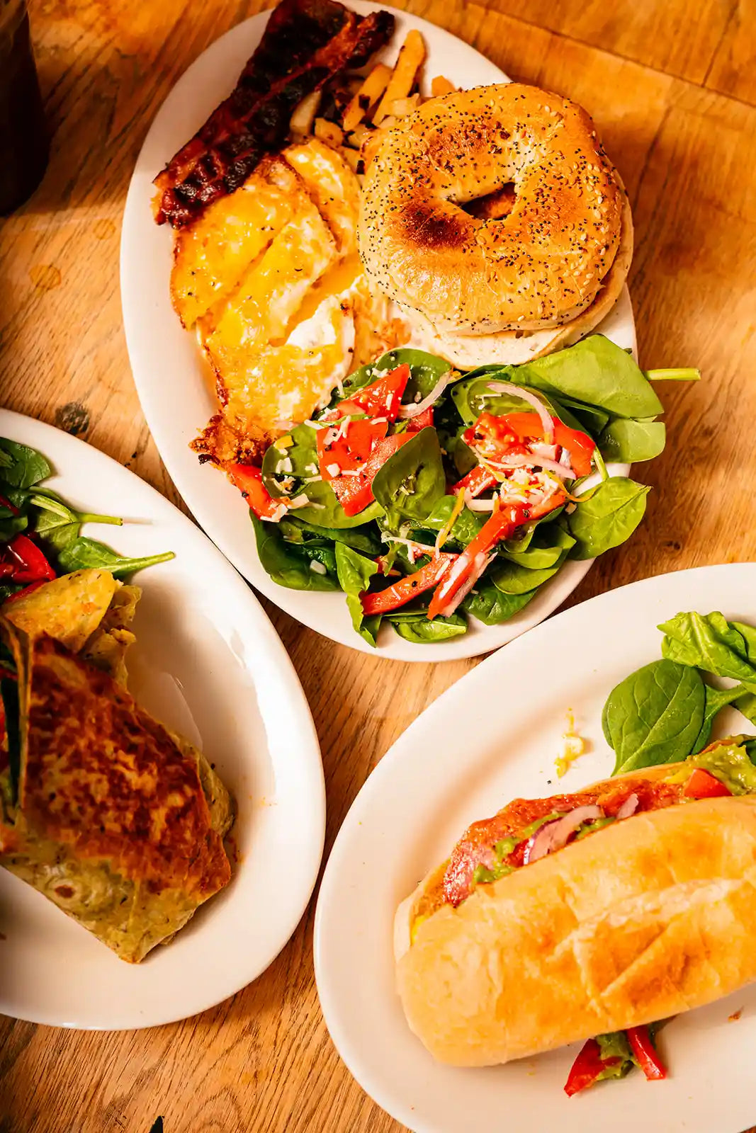 Assorted breakfast dishes including a bagel with spinach and tomato, an omelette, and a sandwich on a wooden table.