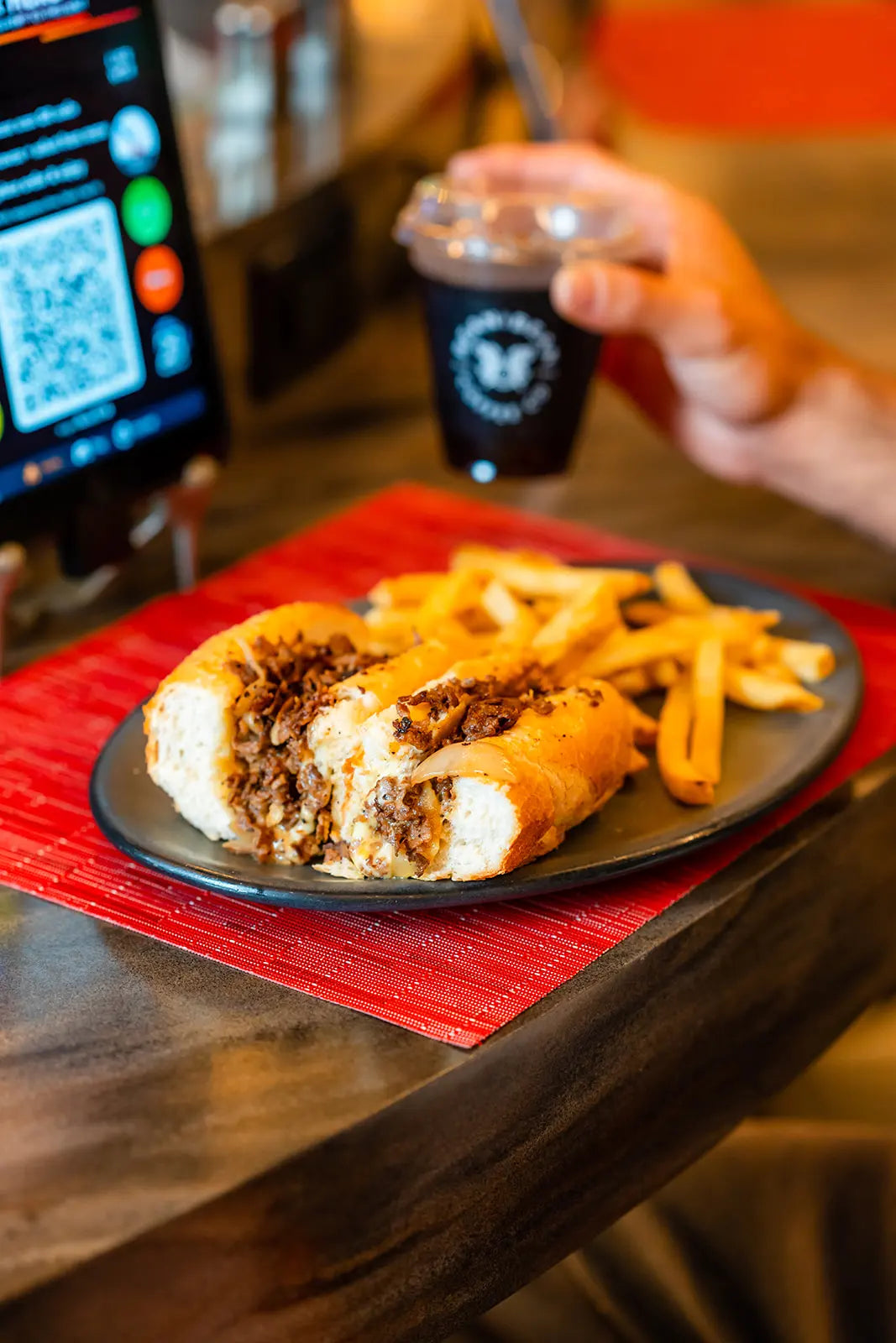 Plate of food with a cheesesteak and fries on a red placemat, with a hand holding a Bean2Bean Cold Brew in the background.