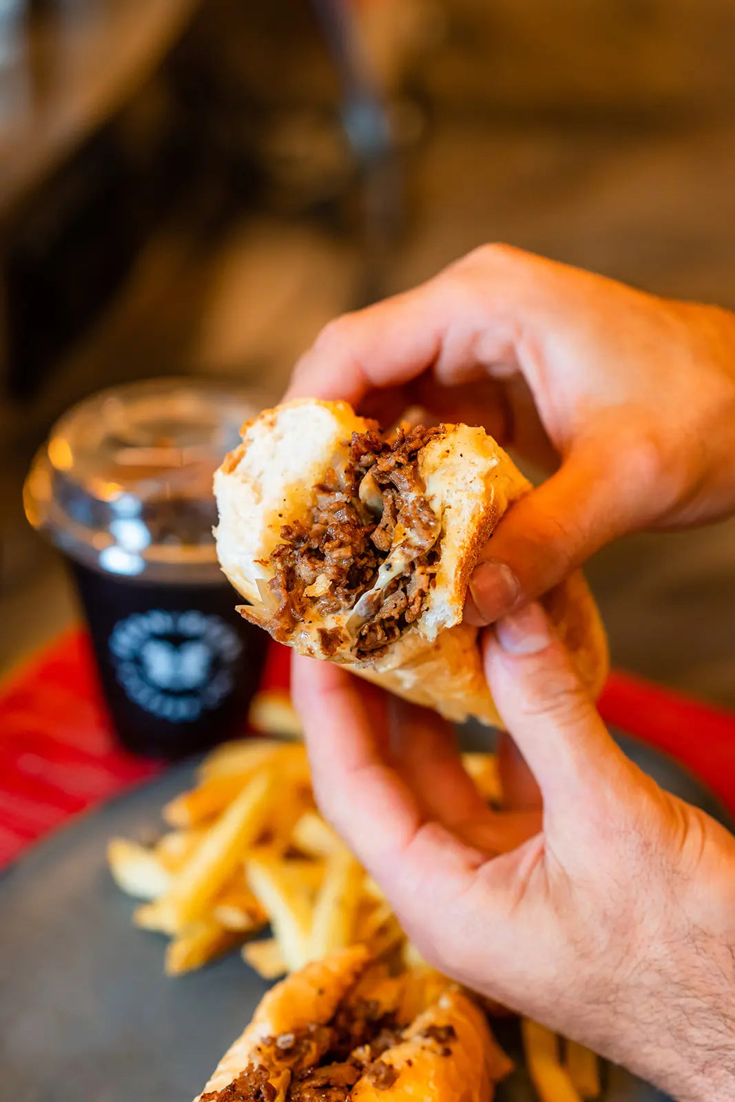Hand holding a cheesesteak cut in half with fries on a table and a Bean2Bean Cold Brew in the background