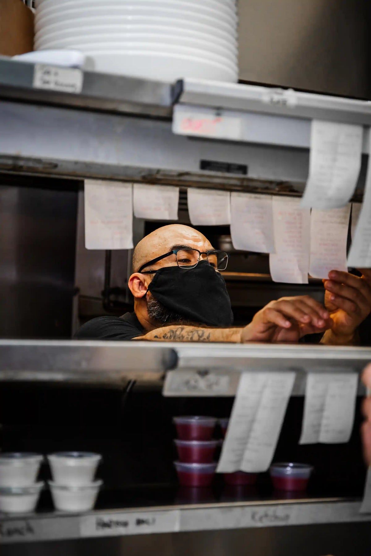 Person wearing a face mask behind a professional kitchen counter with various items including cups and containers.