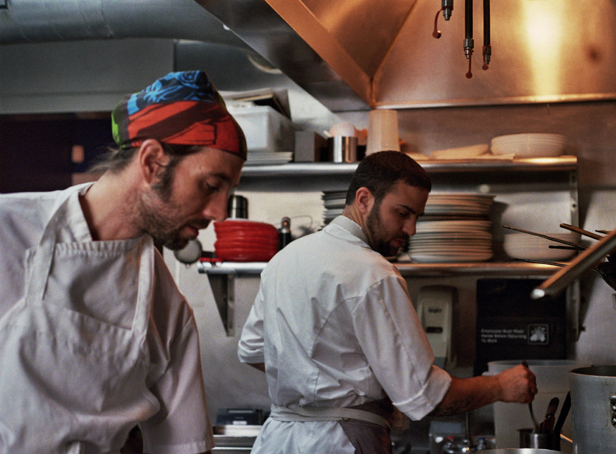 Two chefs working in a kitchen with stainless steel equipment and shelves.