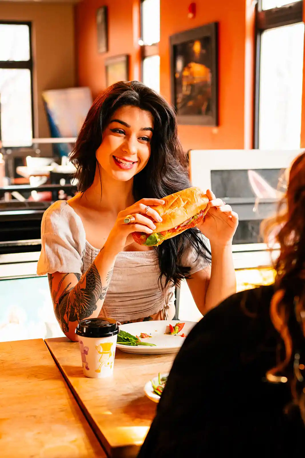 Woman eating a sandwich at a restaurant table with another person partially visible.