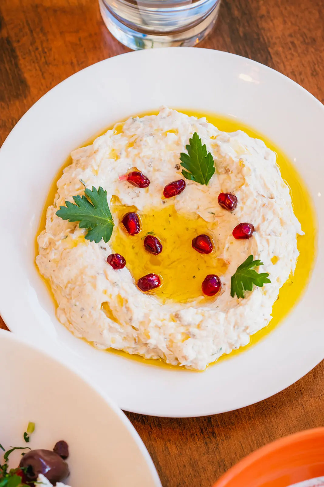 Mutabal with pomegranate and parsley garnish on a white plate on a wooden table.