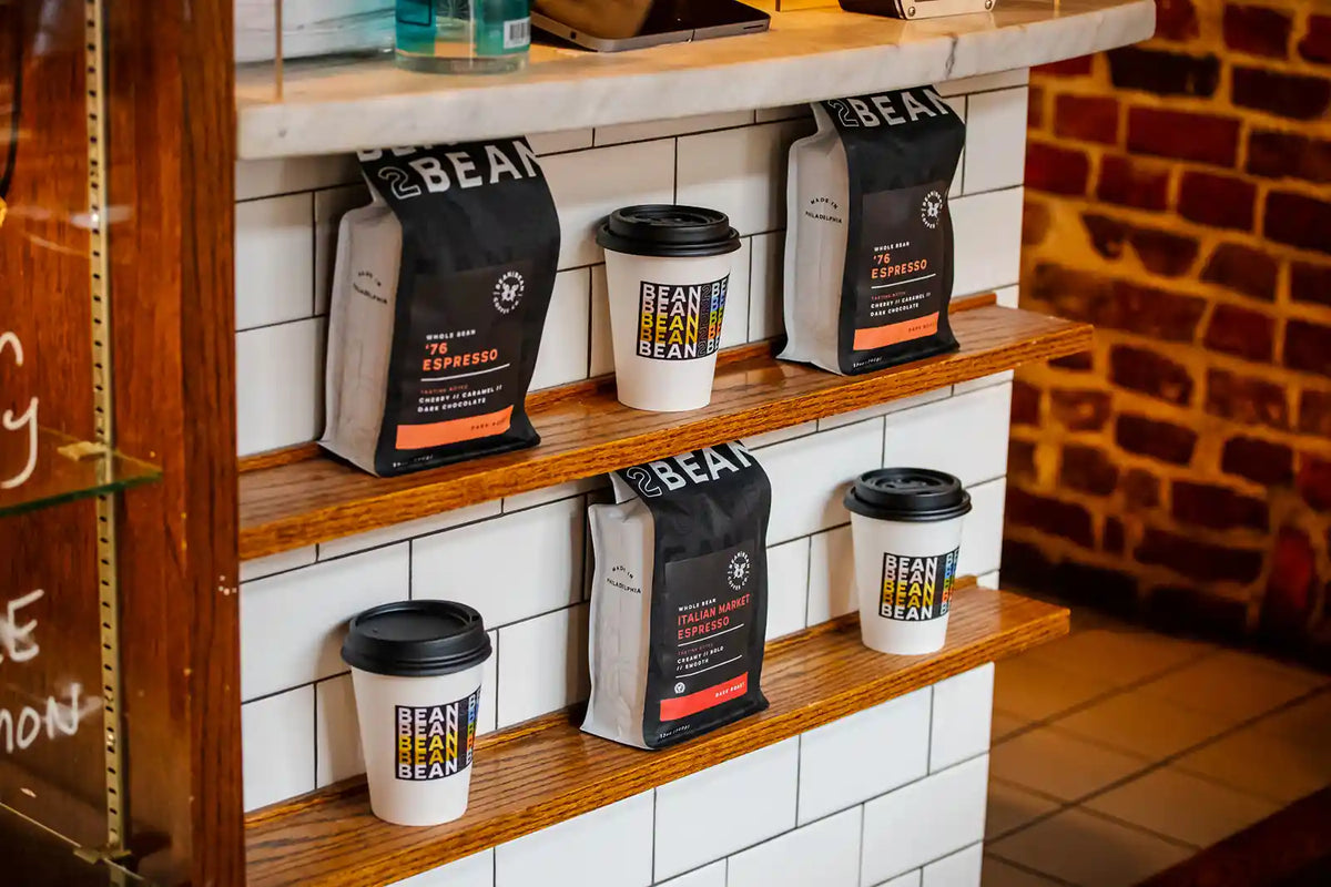 Bean2Bean Coffee bags and cups on a wooden shelf against a tiled wall with a brick accent.