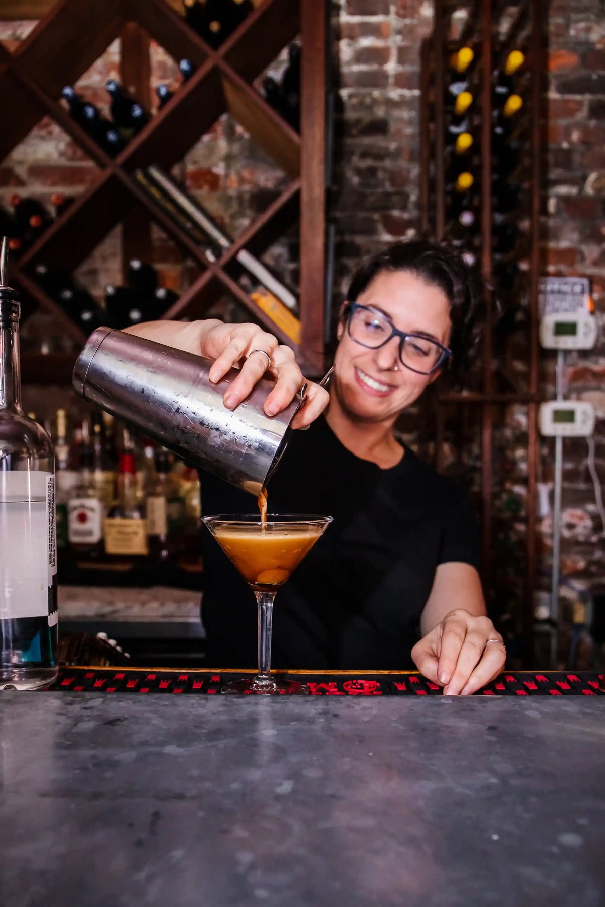 Bartender pouring a drink into a glass with a brick wall and bar setup in the background