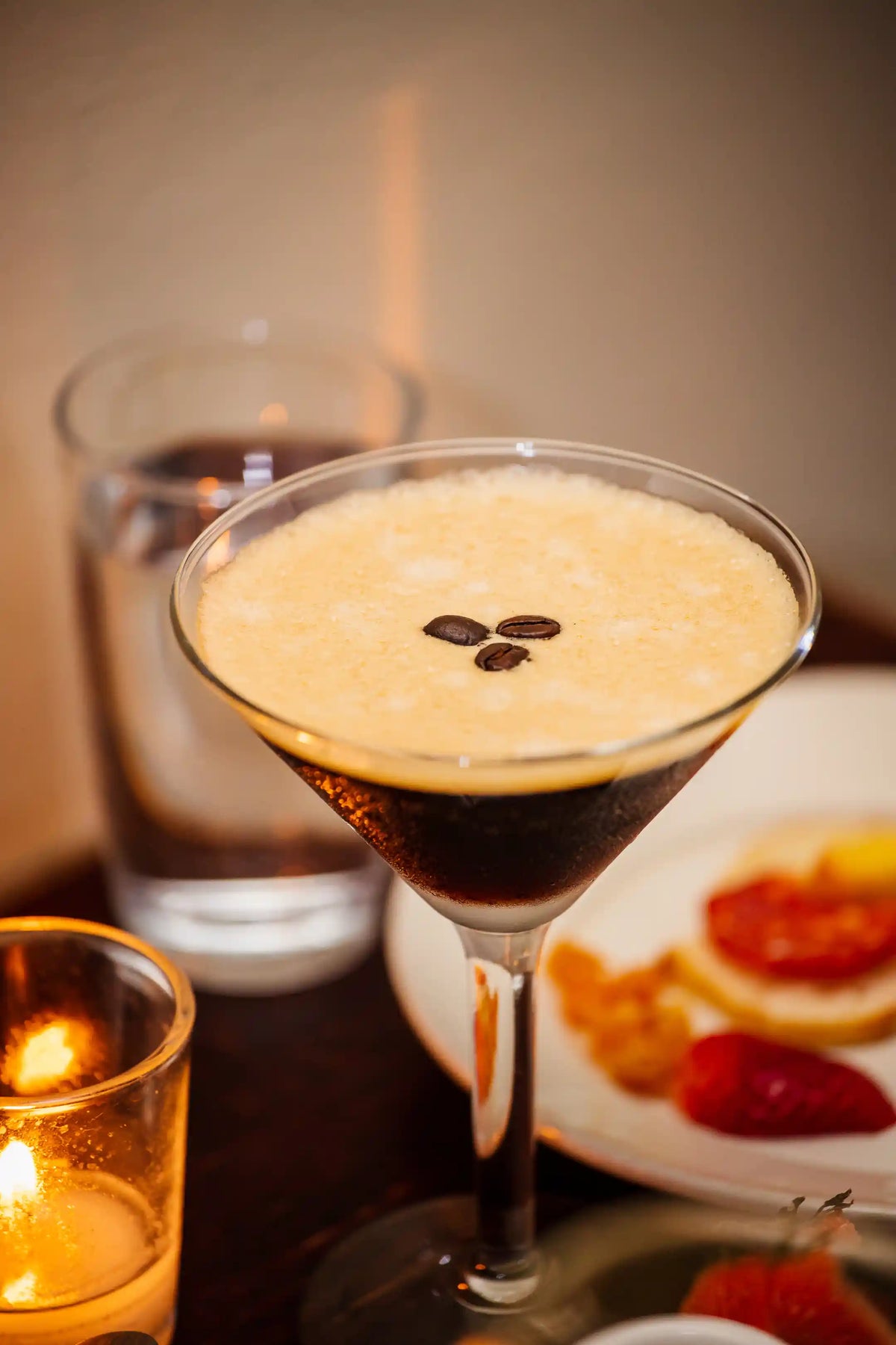 Bean2Bean espresso martini with a creamy top layer and dark liquid base, garnished with coffee beans, on a table with a candle and plate.