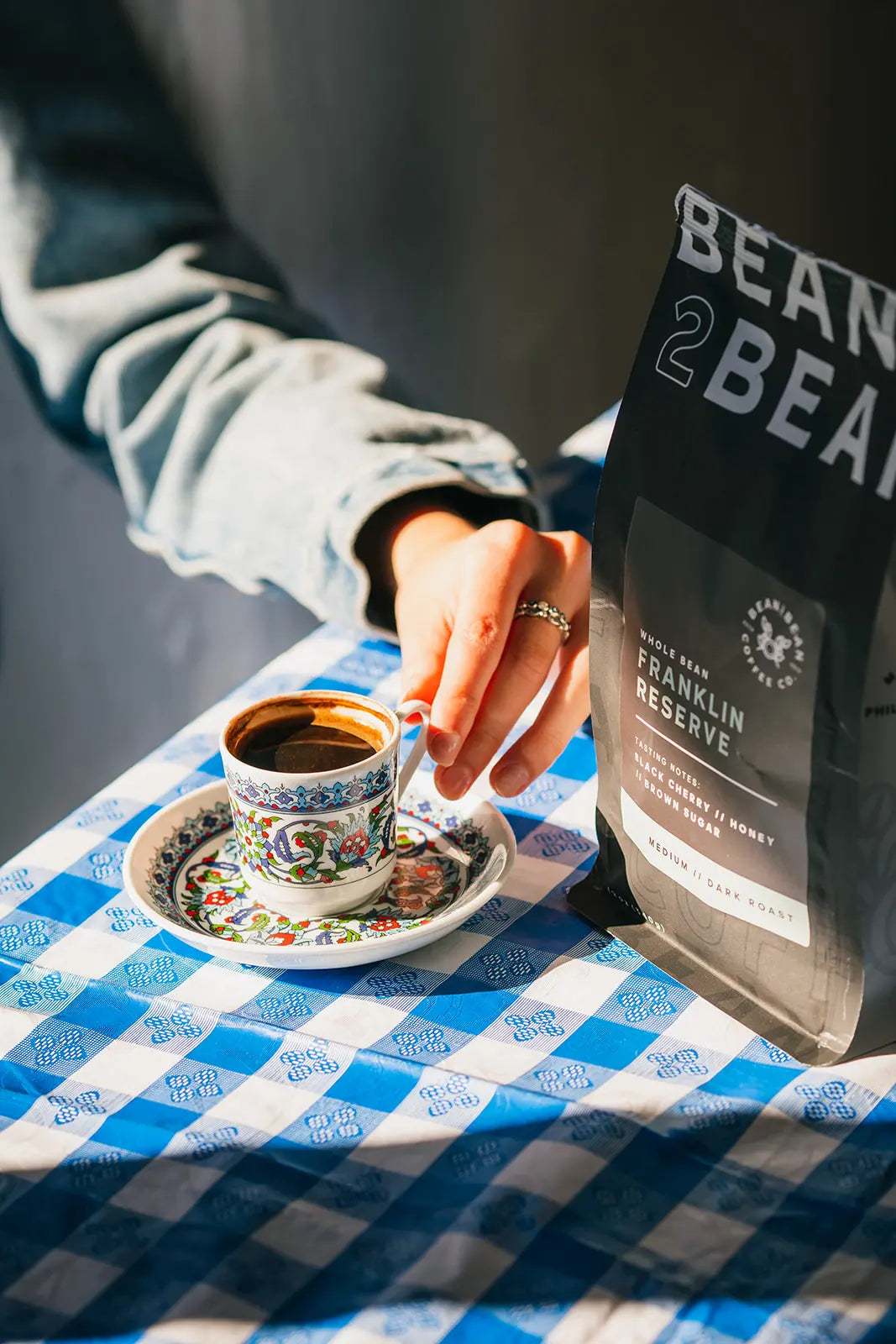 Person holding a cup of coffee with a 'Bean2Bean' coffee bag on a checkered tablecloth.