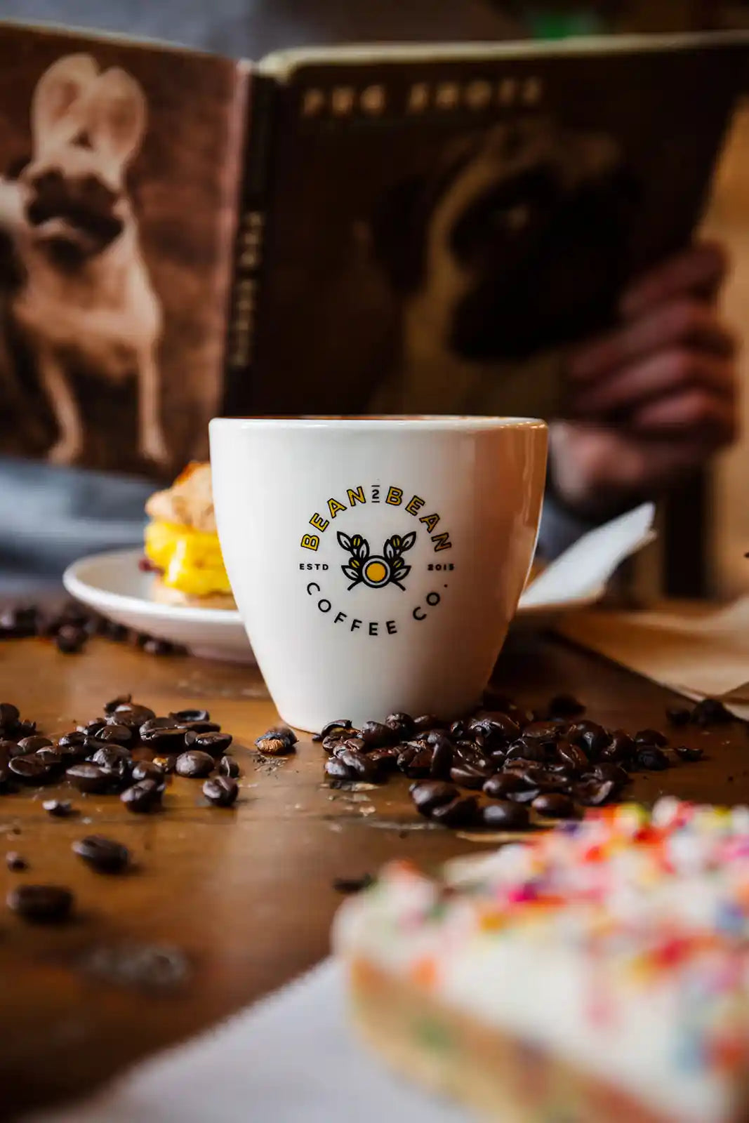 White coffee cup with a Bean2Bean logo on a table with coffee beans and pastries, with a book in the background.