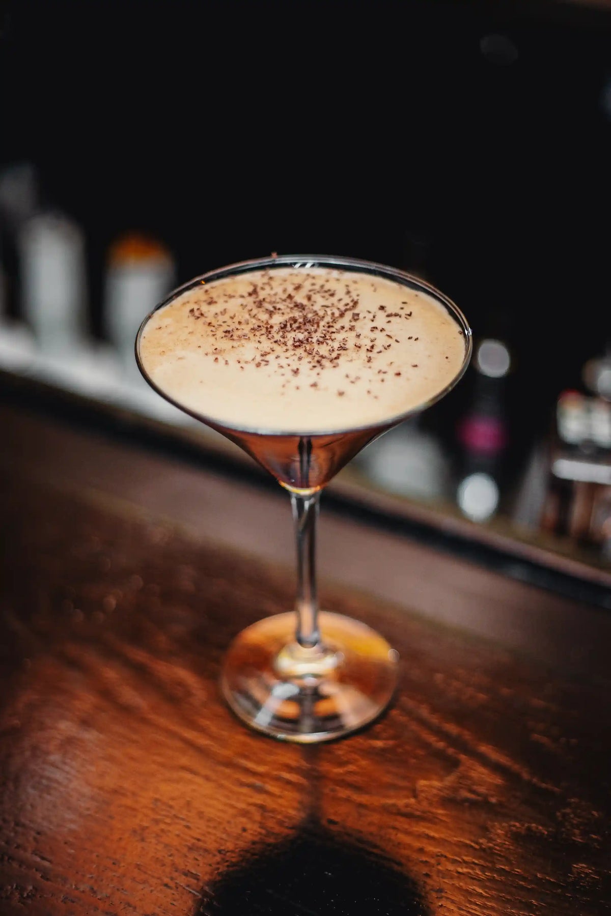 Bean2Bean Espresso martini with a creamy top layer and chocolate shavings in a martini glass on a wooden bar.
