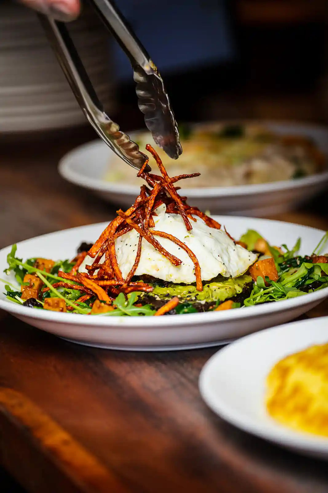Salad with poached egg on a plate, with tongs placing crispy toppings.