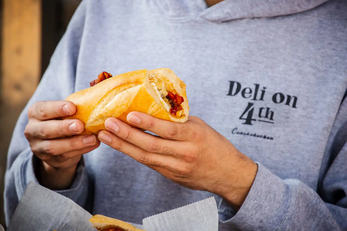 Person in a grey Deli on 4th hoodie sweatshirt holding a breakfast longroll sandwich