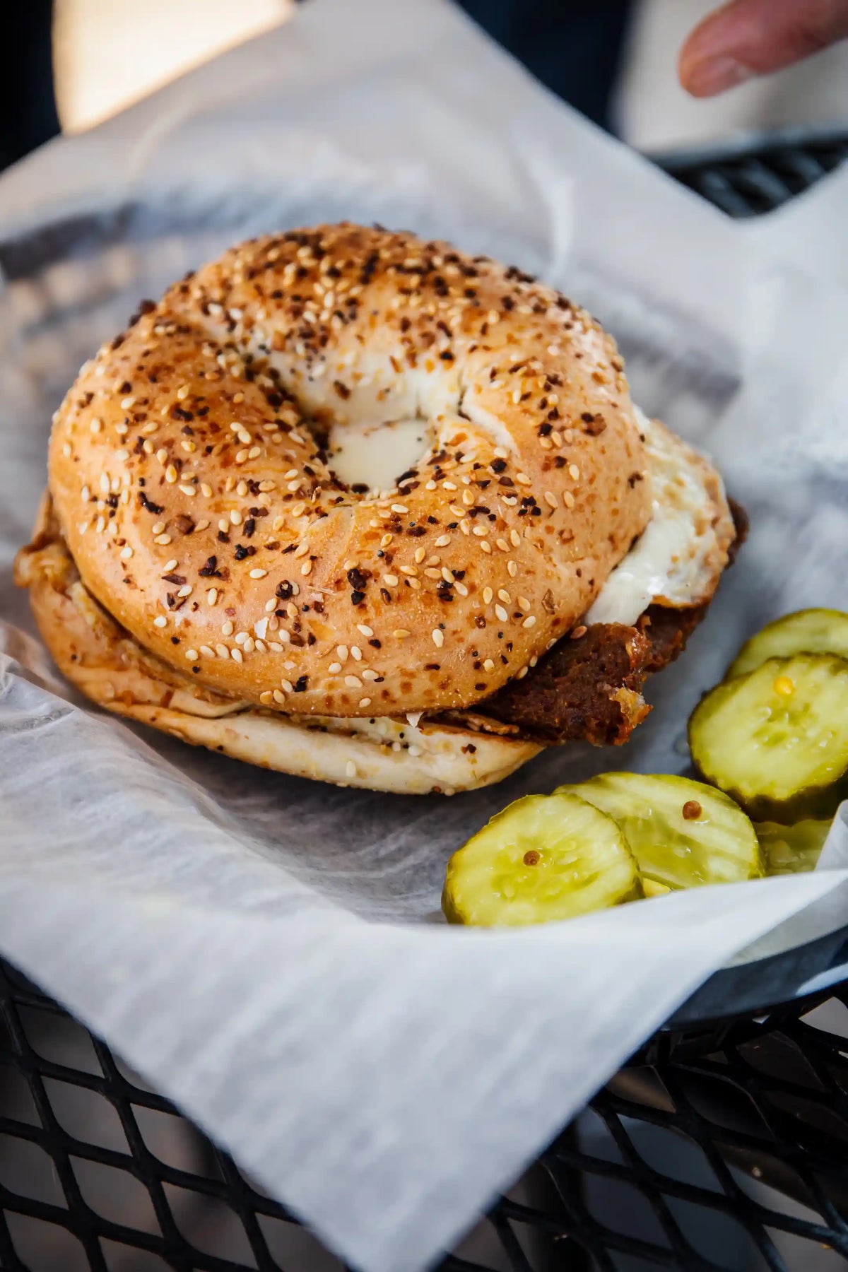 Breakfast sandwich in an everything bagel with pickles on the side in a wax paper basket,