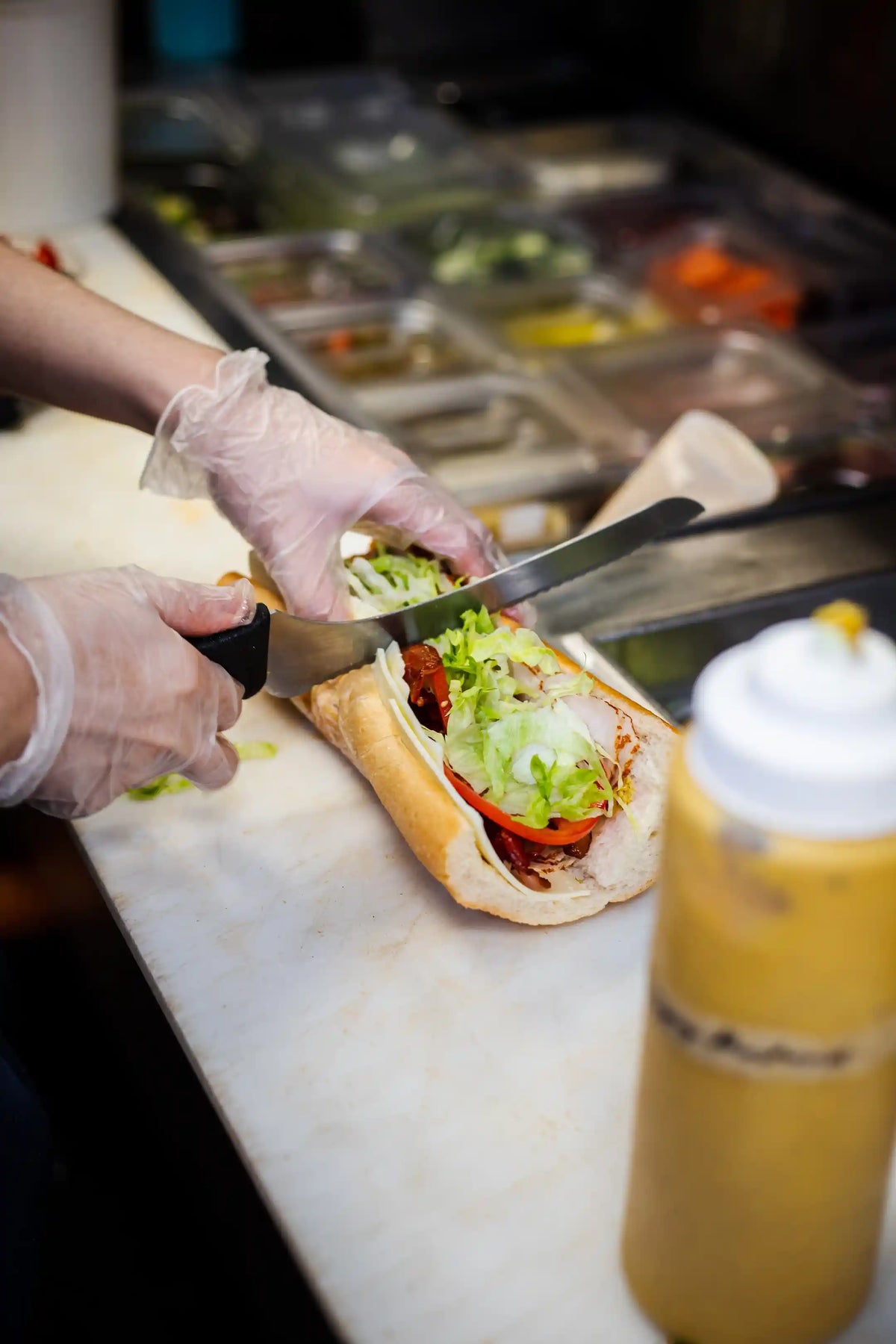 Kitchen employee or chef with gloves cutting a long hoagie sandwich in half on a prep counter