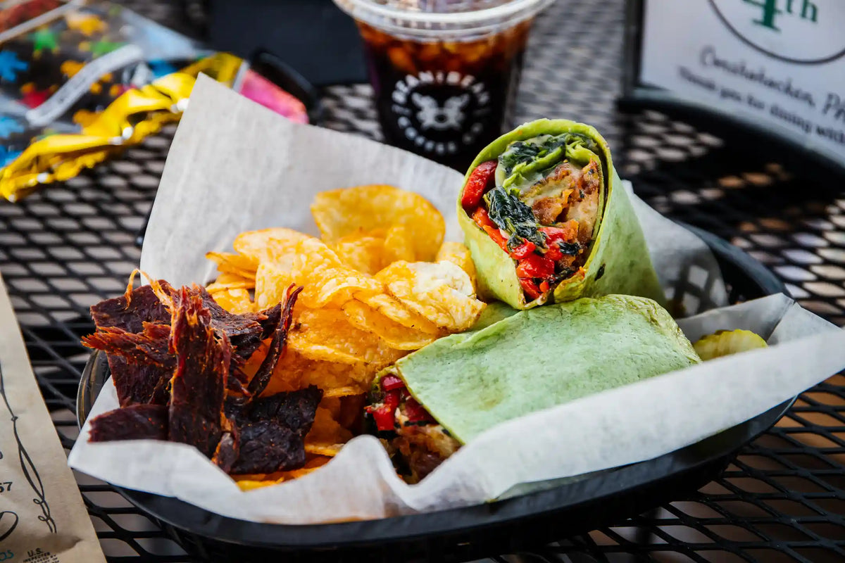 Breakfast wrap with green wrap and vegetables with beef jerkey on the side in basket and a bean2bean coffee in the background blurred