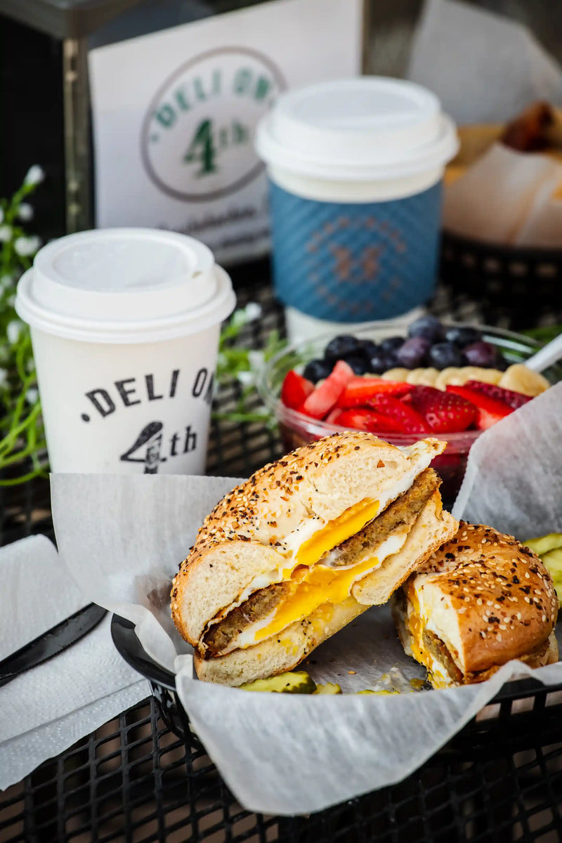 White coffee cup with "Deli on 4th" stamped on it in black on a black table with breakfast sandwich on everything bagel cut in half in basket 