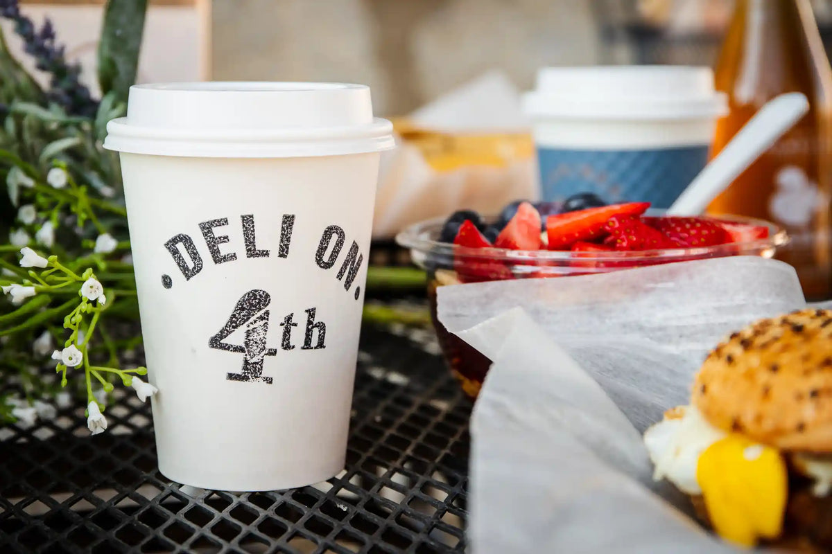 White coffee cup with "Deli on 4th" stamped on it in black on a black table with breakfast sandwich on everything bagel in foreground and fruit cup in background