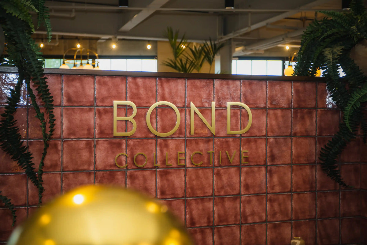 red-ish tile wall with 'BOND COLLECTIVE' logo in gold lettering, surrounded by plants.
