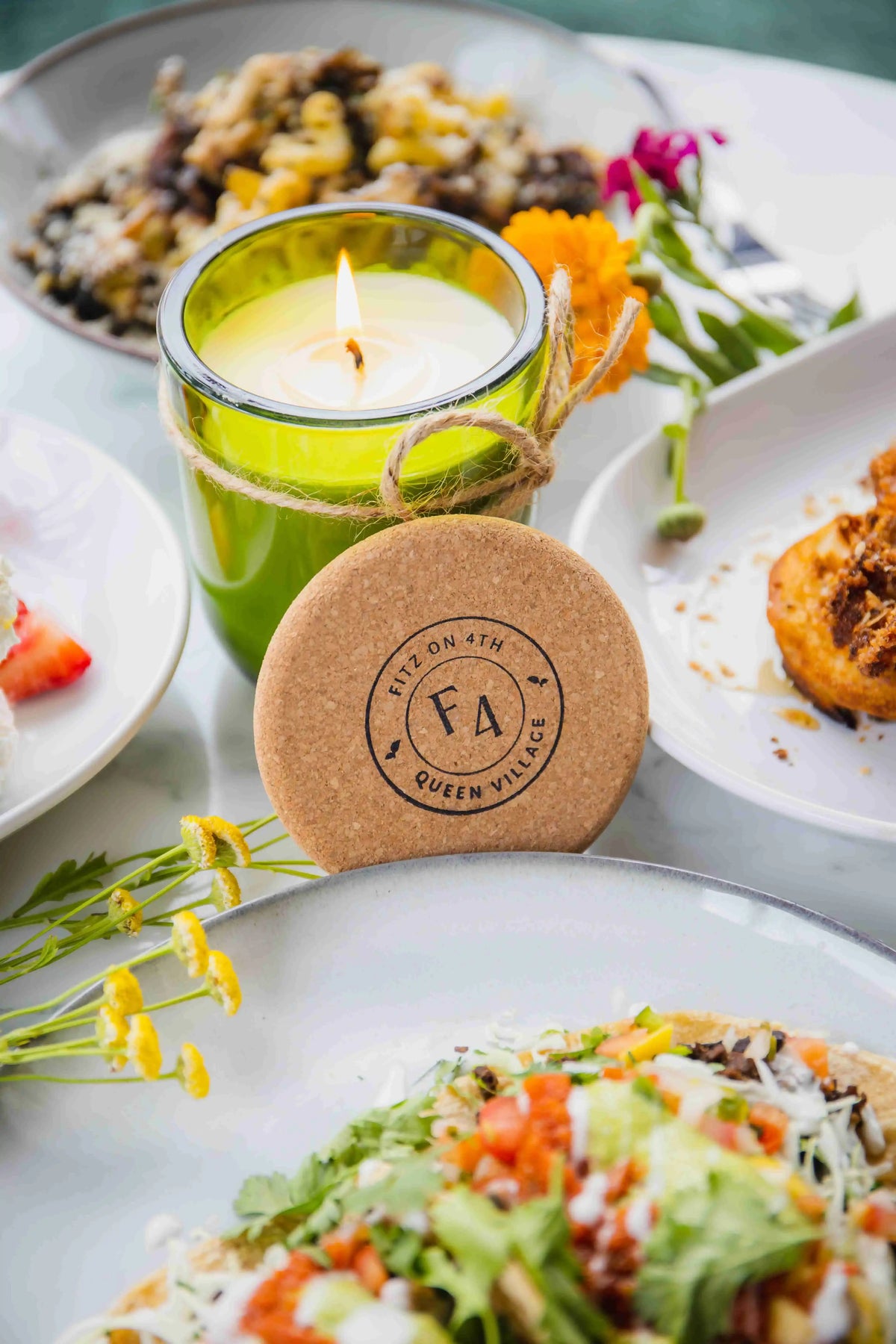 Green glass candle with cork lid leaning against it with "Fitz on 4th" logo on it in black, surrounded by dishes of food