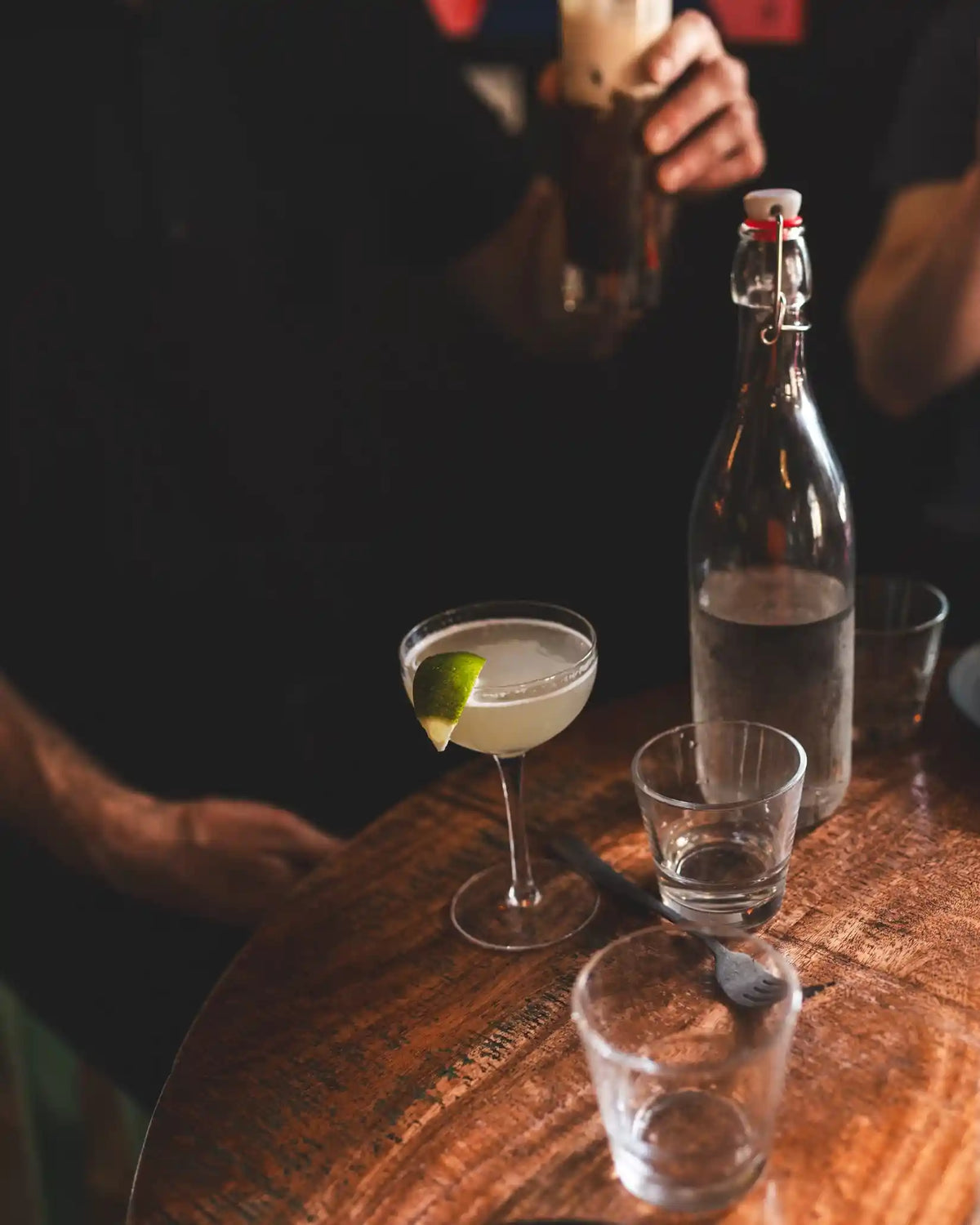 Cocktail with lime garnish on a wooden bar with a bottle and glasses.