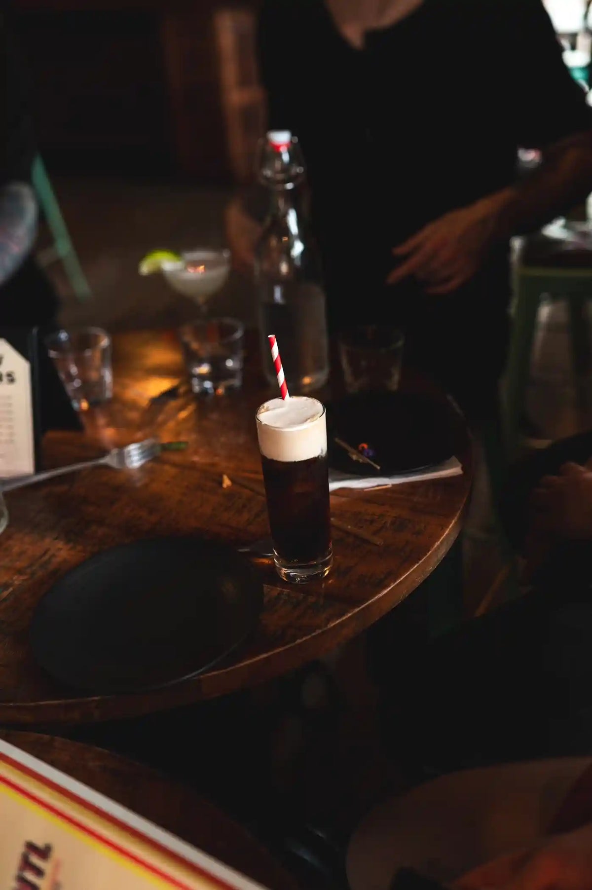 Tall dark cocktail with red and white striped straw on a wooden bar with a bottle and glasses.