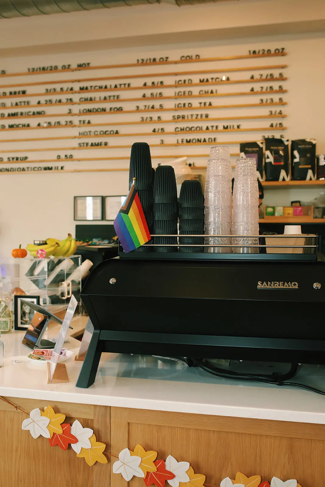 Black espresso machine on a counter with a Pride flag, surrounded by coffee cups and a wall of coffee menu items.