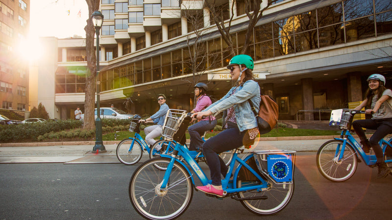 People riding electric Indego bikes on a city street with buildings in the background