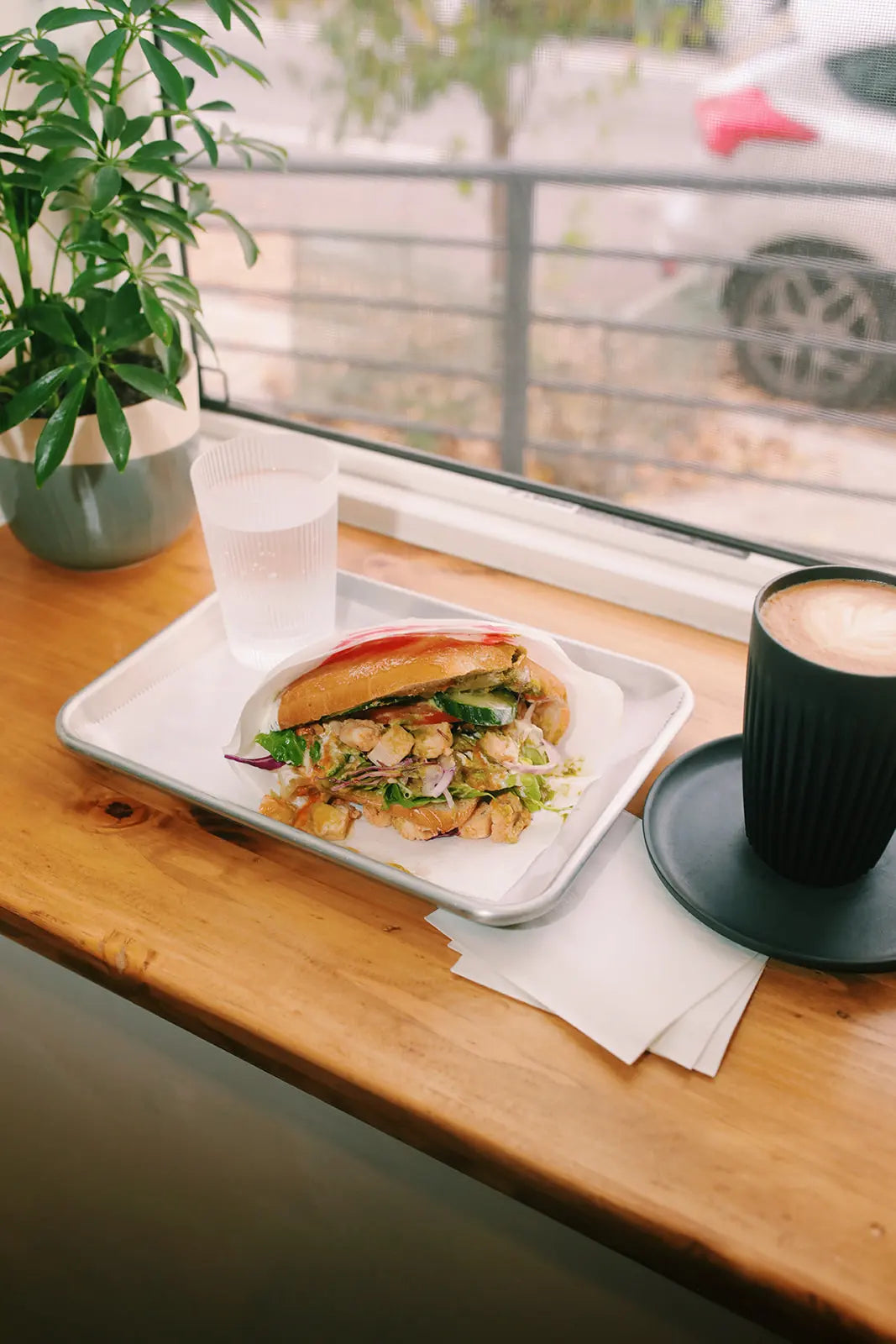 Plated chicken sandwich and cup of Bean2Bean coffee on a wooden table with a window view.