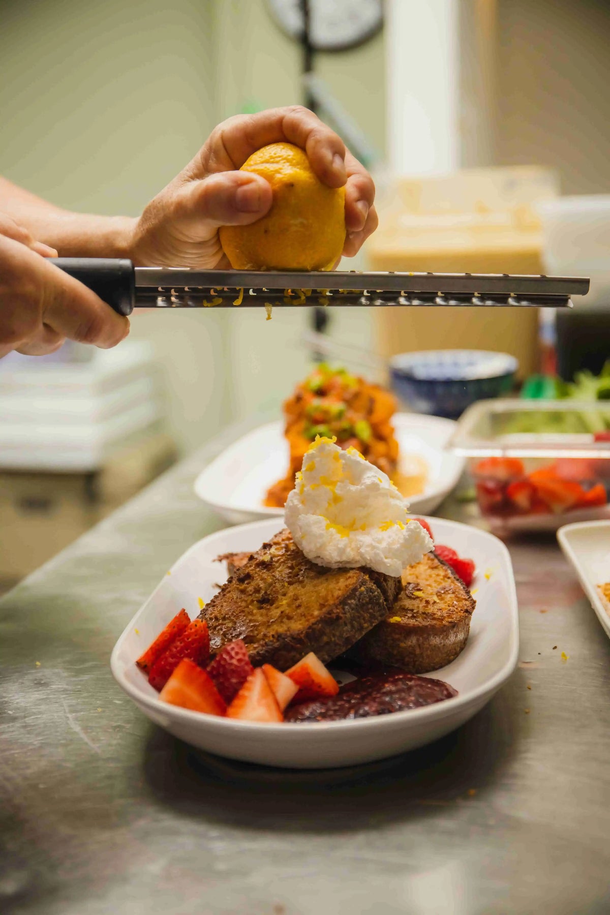 Chef zesting a lemon overtop of a french toast dish on a white plate in kitchen