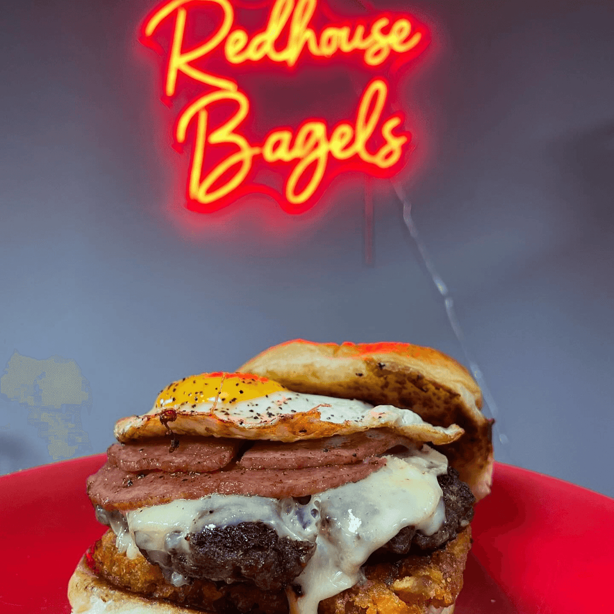 Stacked fried egg and candian bacon burger on a red plate with 'Redhouse Bagels' neon sign in the background