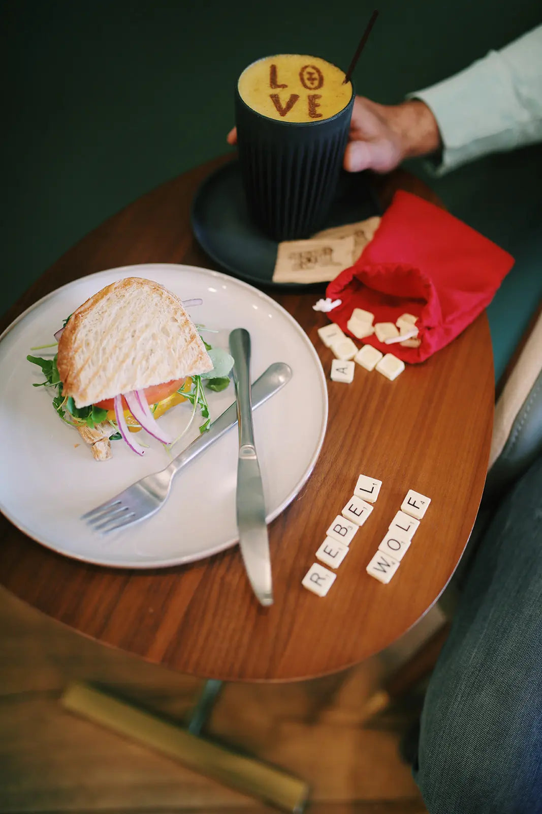 Plate with a sandwich, cup with 'LOVE' design, and scattered letters on a wooden table.