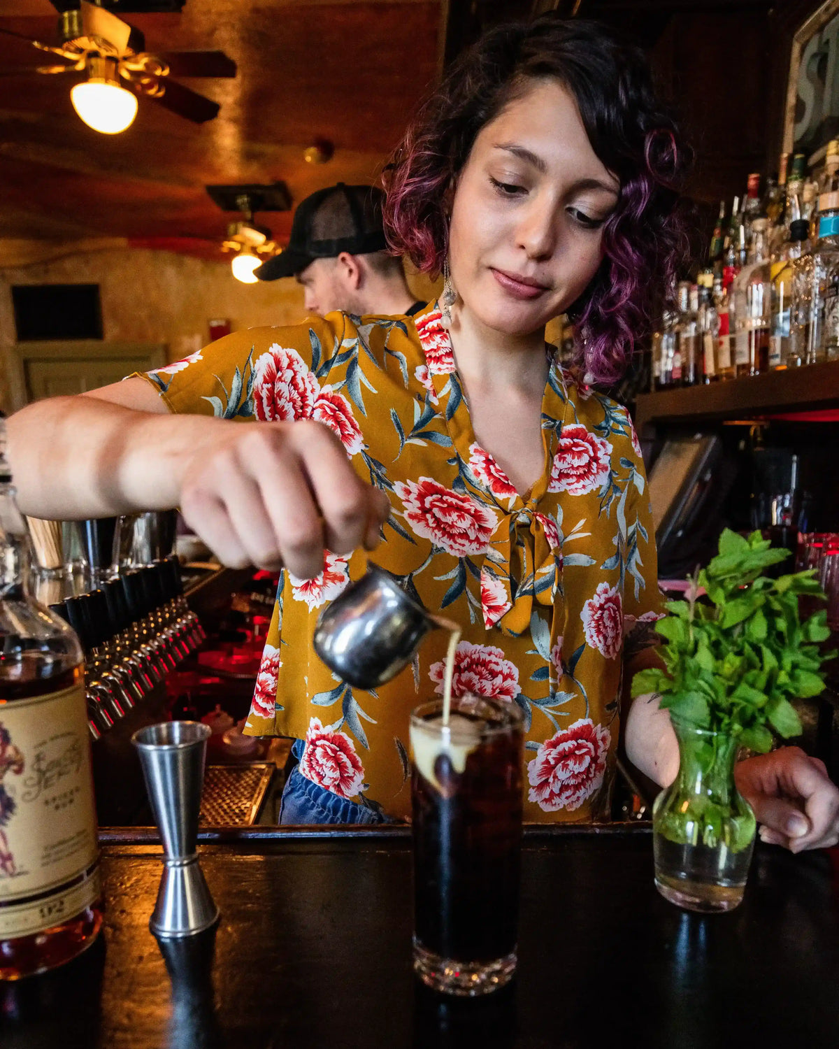 Barista preparing a drink with a floral-patterned shirt in a bar setting.