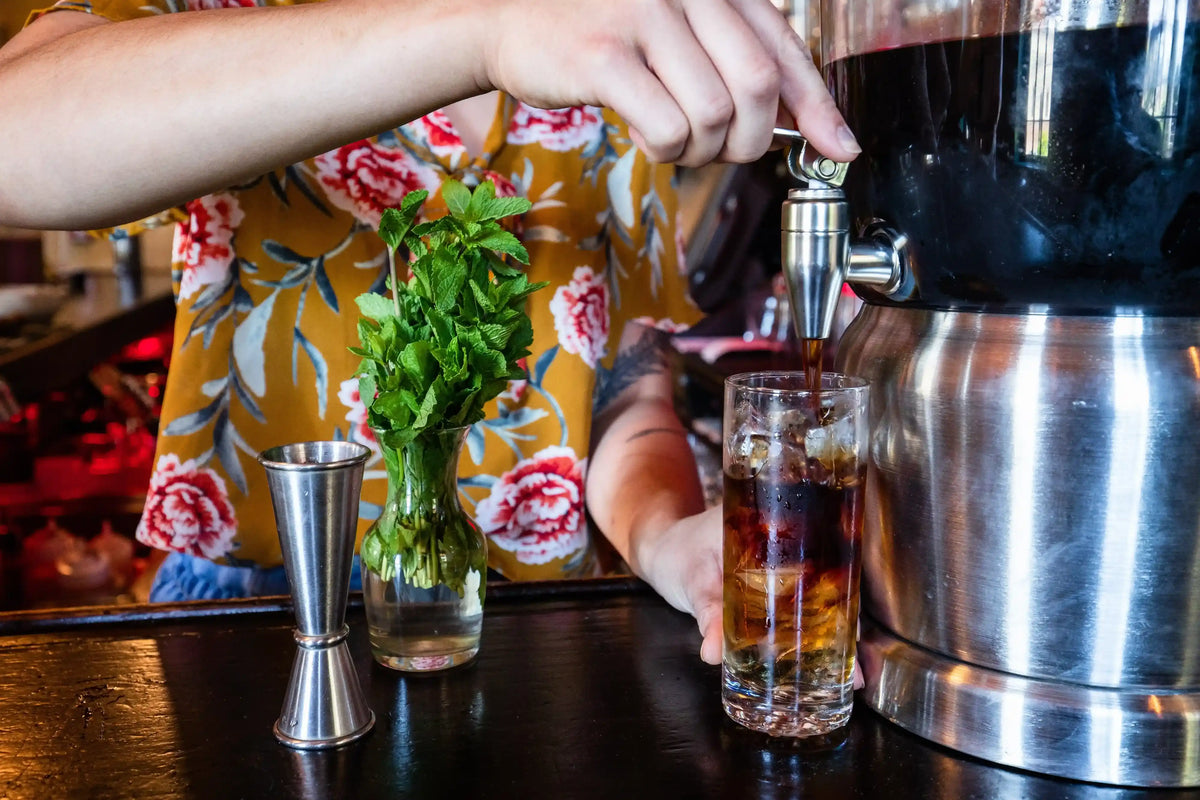 Person pouring a drink from a cold brew tap into a glass with a floral-patterned shirt in the background.