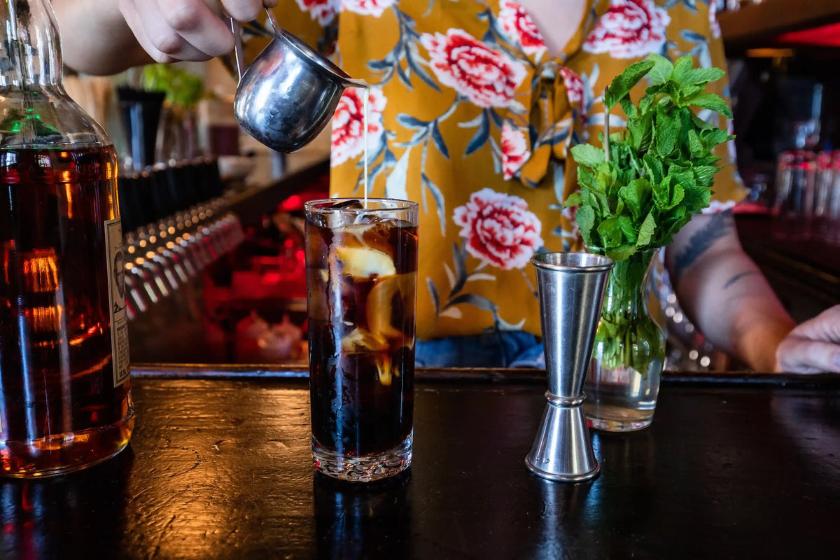 Bartender preparing a drink with a bottle of liquor, glass with ice and liquid, and mint leaves on a bar.