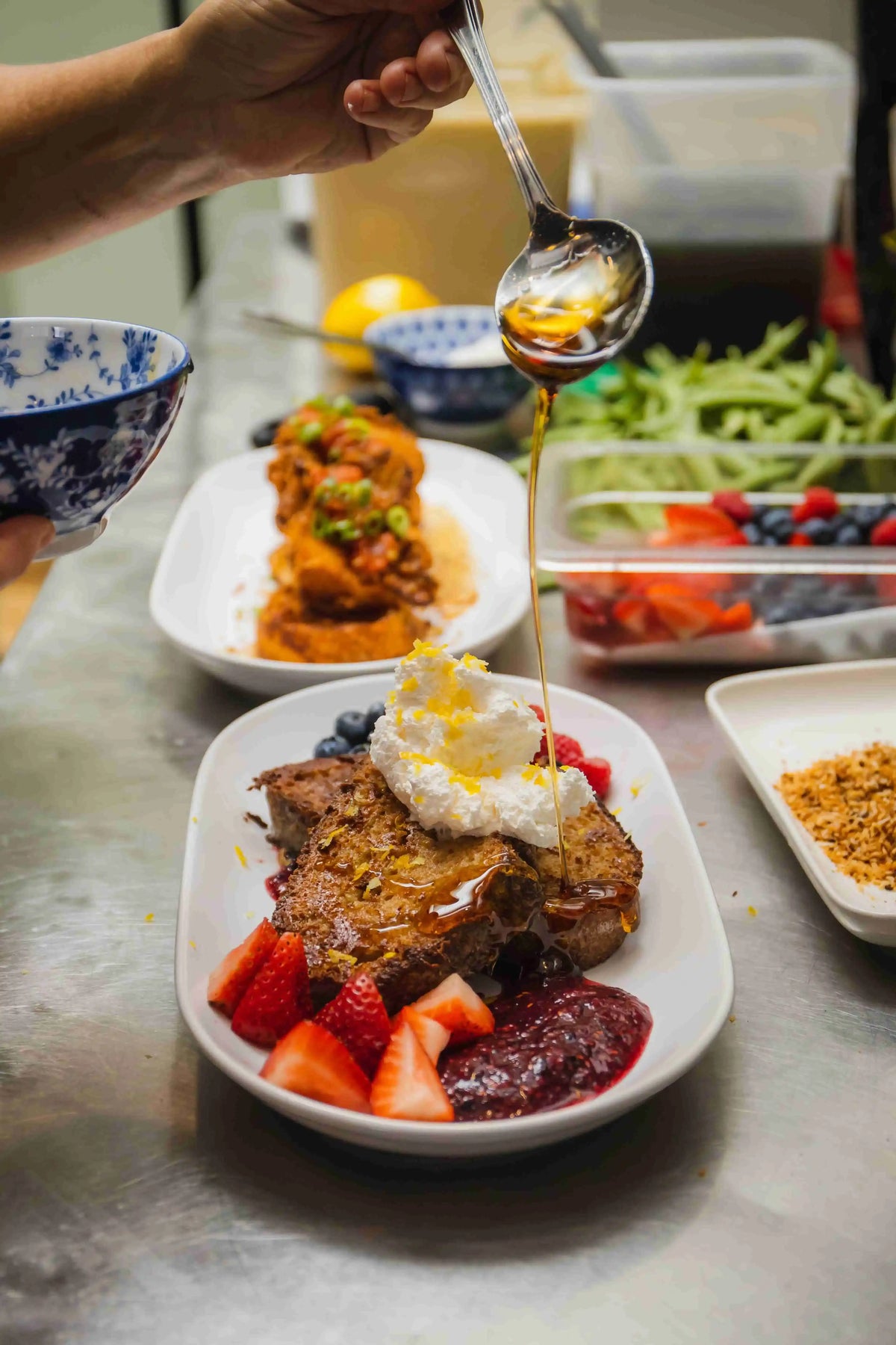 Chef drizzling syrup overtop of a french toast dish on a white plate in kitchen with other food dishes blurred in background