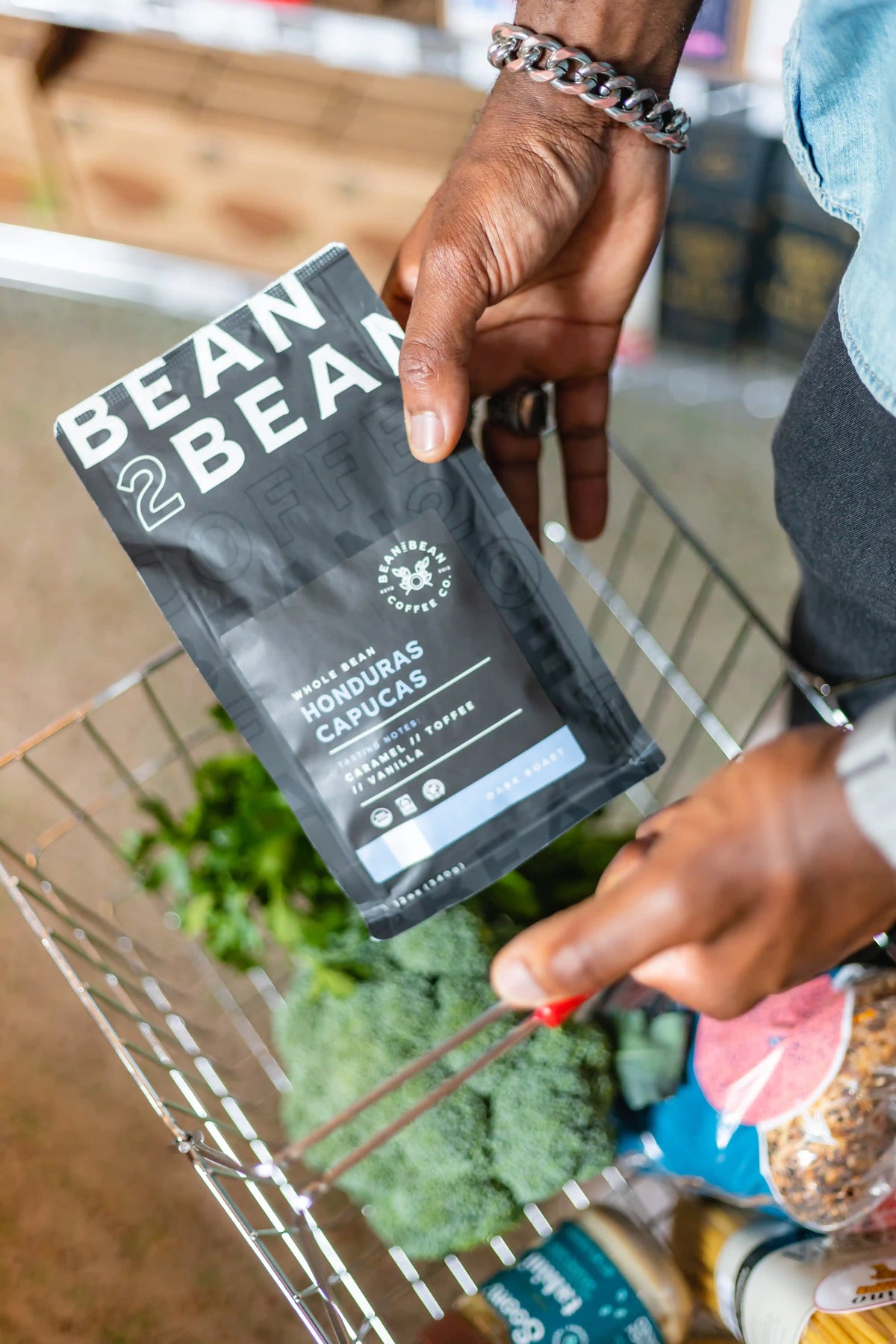 Person holding a 'Bean2Bean' Honduras Capucas coffee package in a shopping cart with groceries.