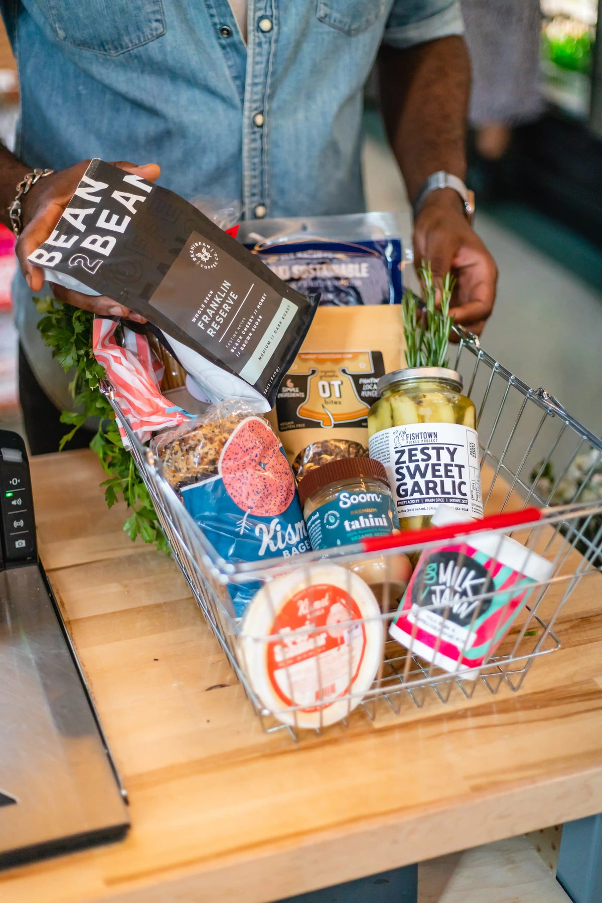 Person holding a shopping basket filled with various food items on a wooden counter.