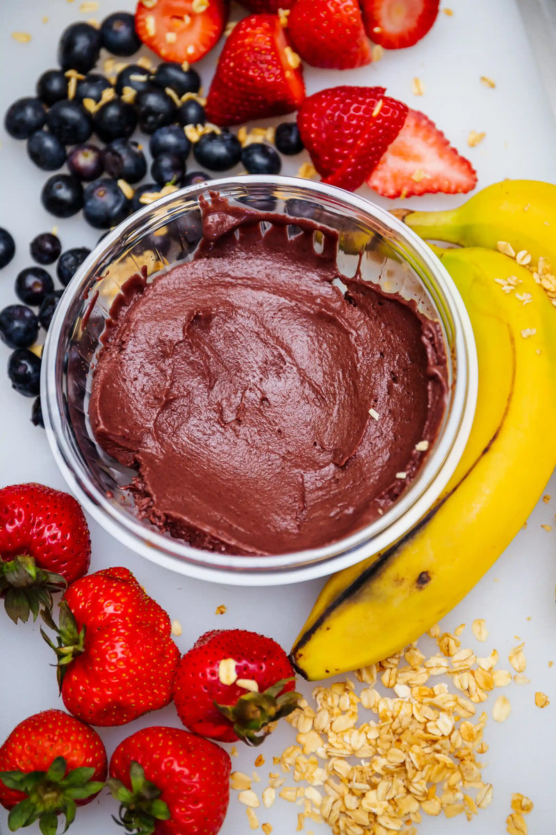 Container of chocolate mouse surrounded by strawberries, bananas, and blueberries on a granie background
