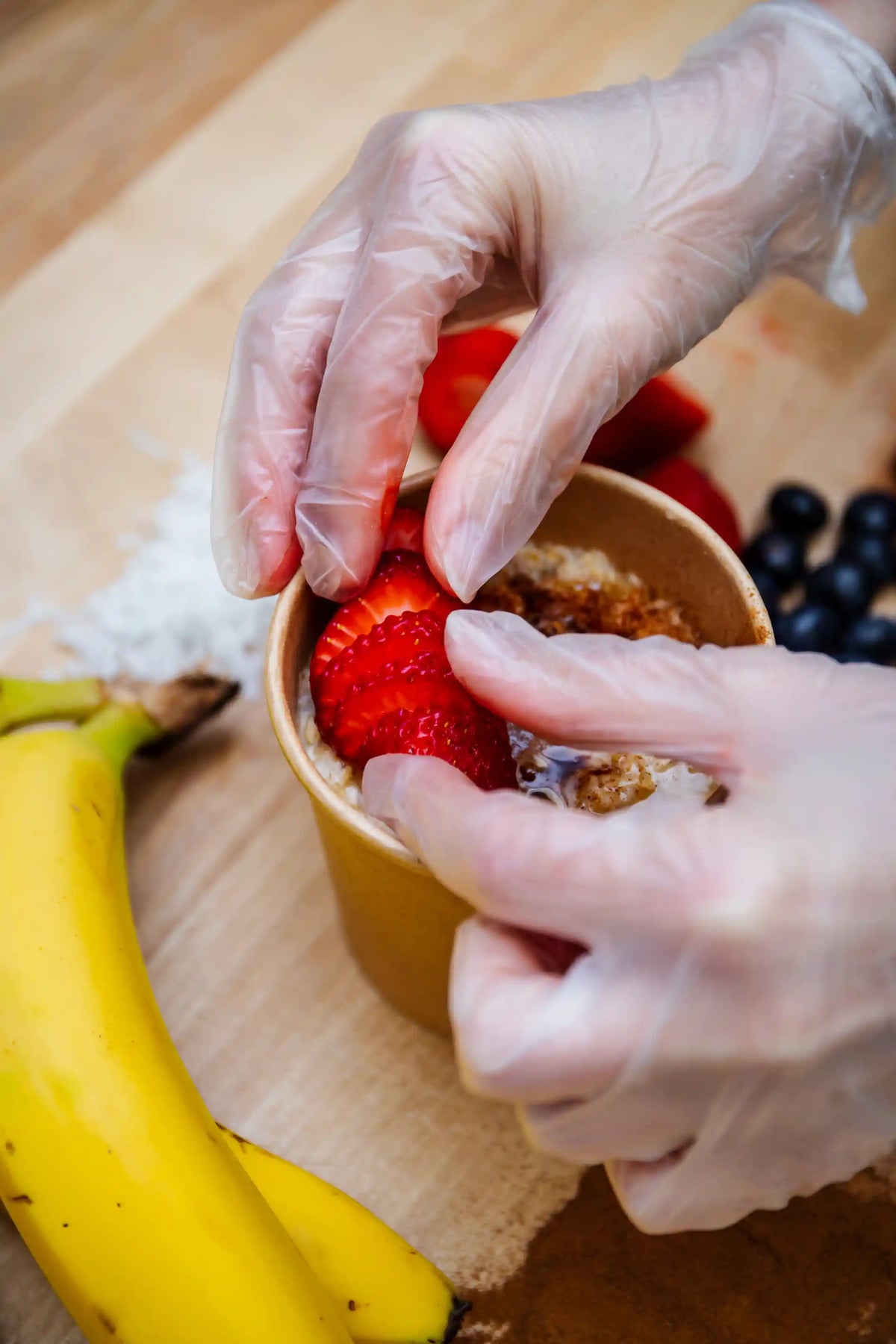 Person preparing an oatmeal bowl with strawberries, bananas, and blueberries on a wooden surface.