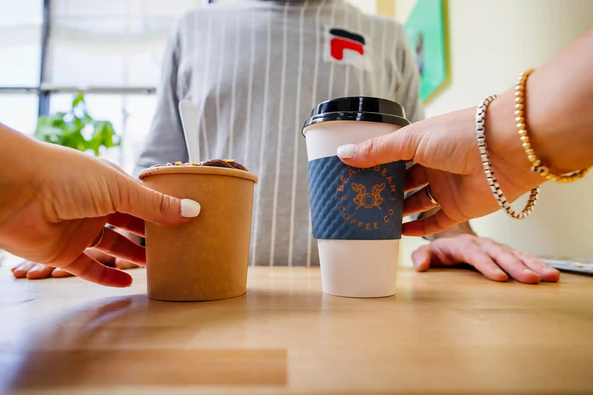 Two hands serving a fruit bowl and a hot Bean2Bean Coffee on a wooden countertop
