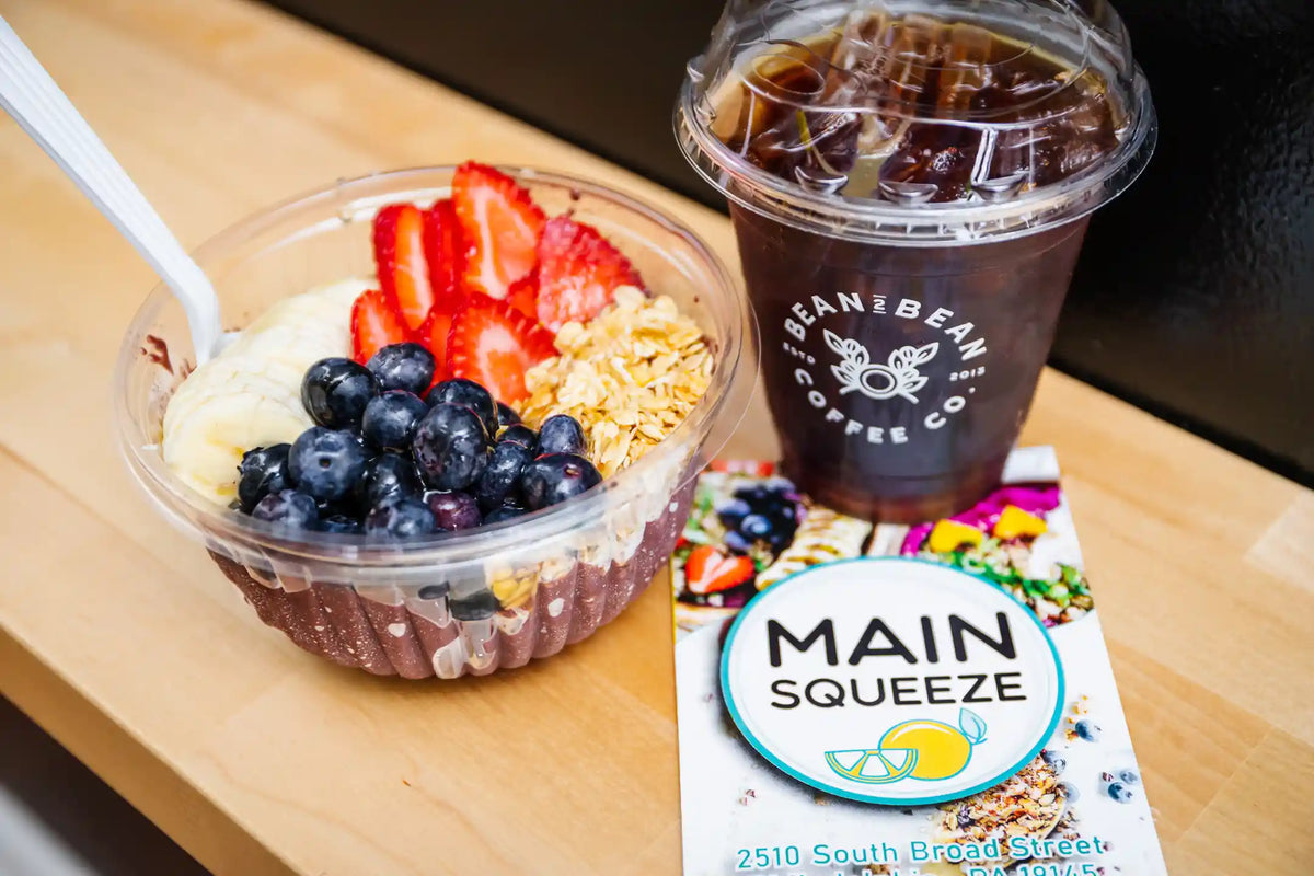 An acai bowl with berries and oats and a cup of Bean2Bean Cold Brew on a wooden table, featuring 'Main Squeeze' menu