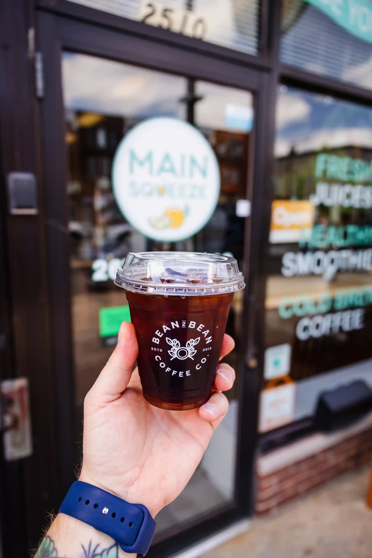 Hand holding a Bean2Bean Cold Brew coffee cup in front of a store with 'Main Squeeze ' sign.
