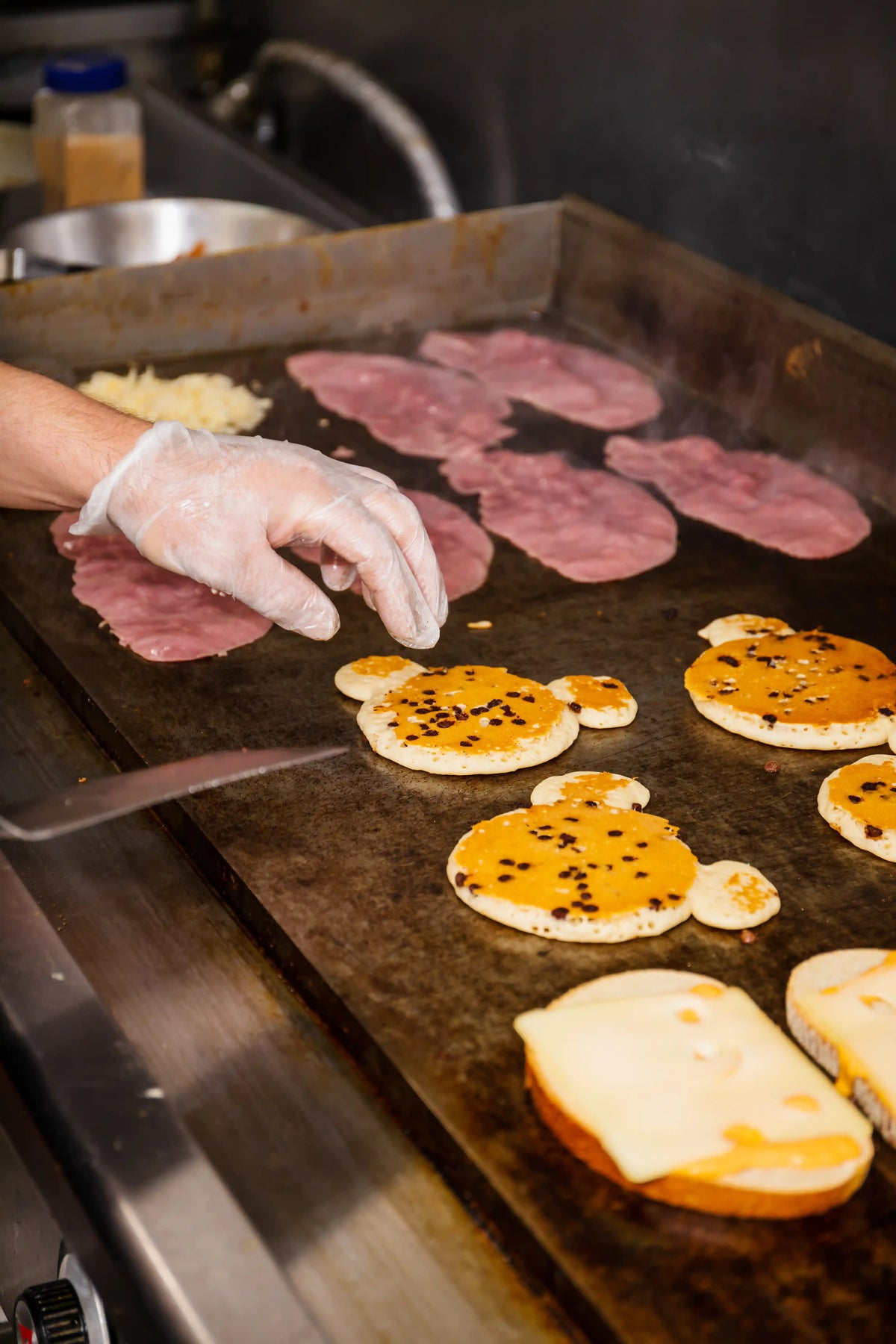Person preparing food on a grill with various ingredients including ham and bread.