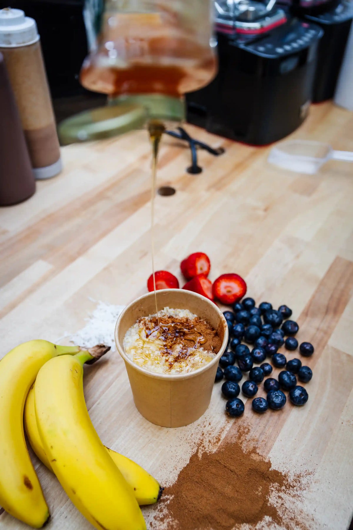 Honey being drizzled into a cup of oats with toppings on a wooden table with bananas, blueberries, and strawberries.
