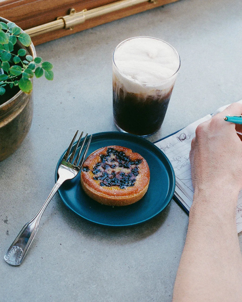 Blue plate with a pastry and an iced latte in a glass on a concrete surface, with a hand holding a pen.