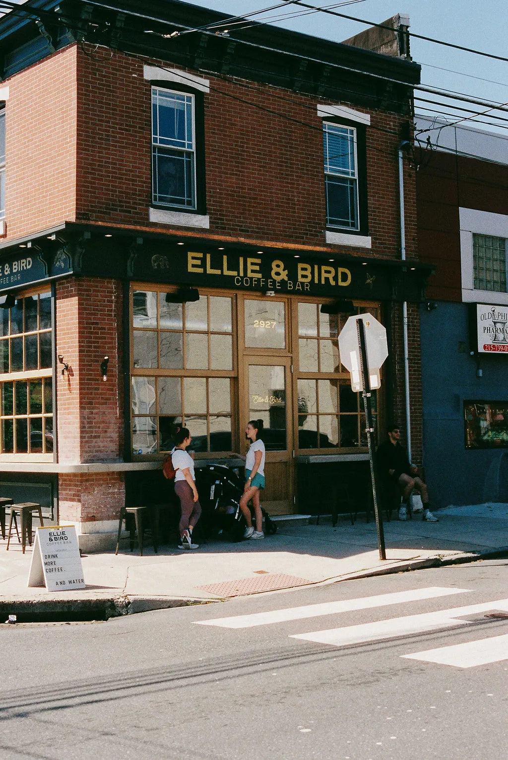 Two people walking past a brick building with 'Ellie & Bird' sign on a city street.